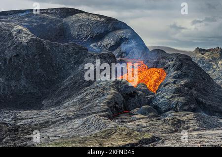 Sprudelnde Lava fließt aus einem aktiven vulkanischen Krater. Landschaft in Island auf der Halbinsel Reykjanes. Krater mit gekühltem und flüssigem Magmagestein. Wolken Stockfoto