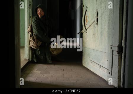 Ein schmutziger Mann in einer Kapuzenjacke sitzt neben der Tür in einem großen dunklen Korridor, hält eine Waffe in den Händen und schaut in den Raum. Das Konzept eines Überlebenden A Stockfoto