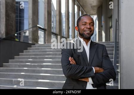 Porträt des erfolgreichen afroamerikanischen Geschäftsmannes Investor, reifer Chef mit gekreuzten Armen lächelnd und Blick auf die Kamera, leitender Mann vor dem Bürogebäude in der Nähe der Treppe. Stockfoto