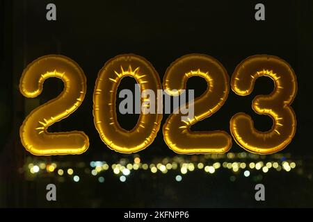 Gold aufblasbare Folienballons Zahlen 2023 auf dem Fenster vor dem Hintergrund der Nachtstadt, in Neonlicht. Neujahr 2023, weihnachten. Wir feiern Weihnachten und Neujahr 2023. Stockfoto