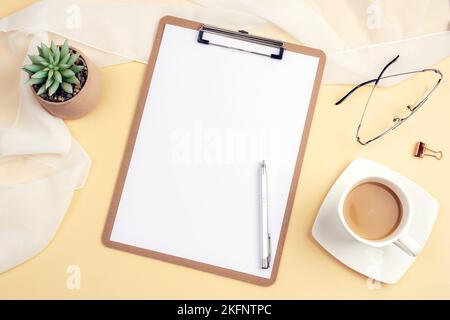 Schreibtisch mit leerem Klemmbrett, Kaffeetasse, Stift, Brille und Zubehör auf hellbeigem Hintergrund. Draufsicht, flaches Lay, Mockup. Stockfoto