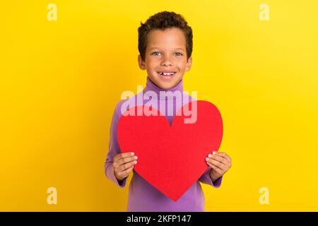 Foto Porträt von charmanten kleinen Schüler Junge halten große rote Herz Papierform tragen trendige violette Kleidung auf gelbem Hintergrund isoliert Stockfoto