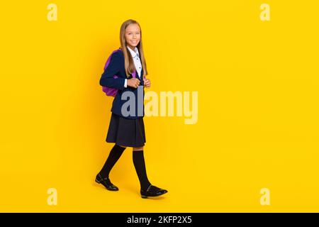 Foto in voller Größe von positiven fröhlichen Mädchen mit geraden Frisur gekleidet blaue Uniform zur Schule gehen isoliert auf gelbem Hintergrund Stockfoto