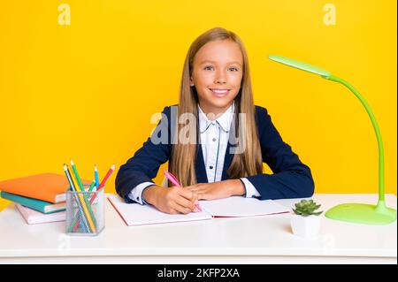 Foto von smart schöne positive Schulmädchen gerade Frisur tragen blauen Blazer sitzen am Schreibtisch schriftlich isoliert auf gelbem Hintergrund Stockfoto