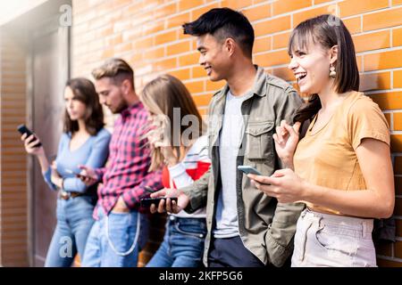 Multikulturelle Freunde lachen mit dem Smartphone an der Wand auf dem Universitätscampus - Junge Menschen süchtig nach mobilen Smartphones - Technologie conce Stockfoto