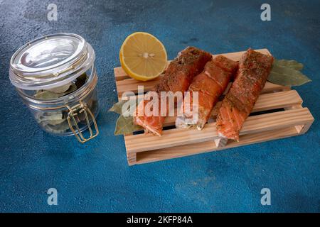 Drei Forellen liegen auf einem Brett auf blauem Hintergrund. Fisch mit Gewürzen bestreut. In der Nähe liegt eine halbe Zitrone und ein Glas Lorbeerblätter. Stockfoto