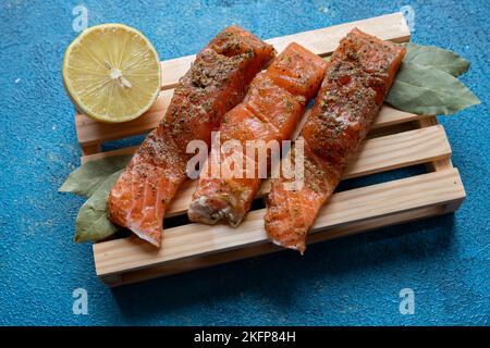 Drei Forellen liegen auf einem Brett auf blauem Hintergrund. Fisch mit Gewürzen bestreut. In der Nähe liegt eine halbe Zitrone und ein Lorbeerblatt. Stockfoto