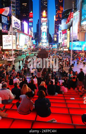 Auf den Stufen und Sitzen des TKTS-Gebäudes versammelt sich eine Menschenmenge, um einen Blick auf die beleuchteten Videotafeln mit Werbung am Times Square, New York, zu werfen Stockfoto