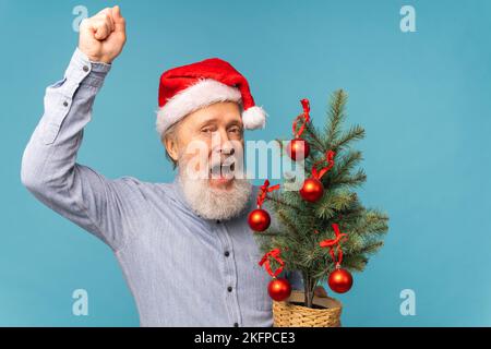 Portrait von fröhlichen, verrückten Emotionen der Weihnachtsmann schaute in die Kamera und hielt einen kleinen weihnachtsbaum auf blauem Hintergrund Stockfoto