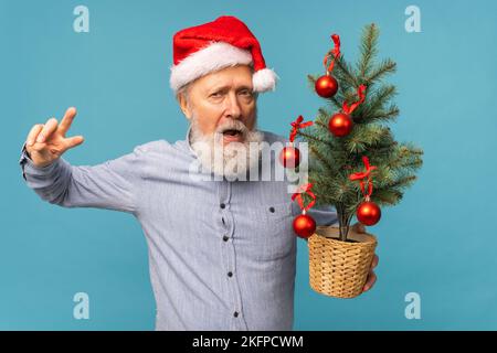 Portrait von fröhlichen, verrückten Emotionen der Weihnachtsmann schaute in die Kamera und hielt einen kleinen weihnachtsbaum auf blauem Hintergrund Stockfoto