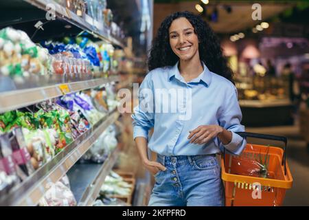 Porträt einer glücklichen hispanischen Frau, die im Supermarkt einkauft, Frau mit einem lächelnden Warenkorb und Blick auf die Kamera, in der Nähe von Regalen mit Produkten in der Lebensmittelabteilung. Stockfoto