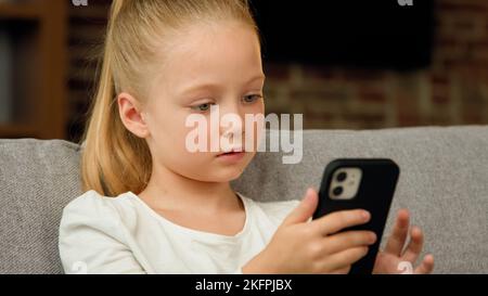Liebenswert kleines Kind Mädchen niedliches Kind sitzt auf gemütliche Couch allein im Wohnzimmer Blick auf Gadget-Bildschirm mit Smartphone-Software-Anwendung spielen Stockfoto