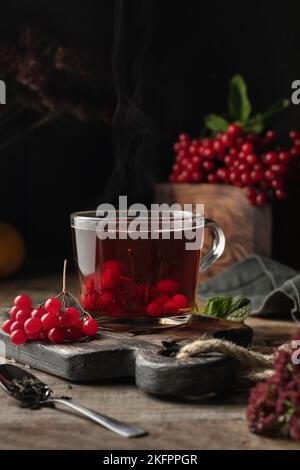 Viburnum-Tee in Glasbecher mit Dampf. Gesundes Getränk der Herbstsaison. Düsteres launiges Stillleben. Vertikale Ausrichtung Stockfoto