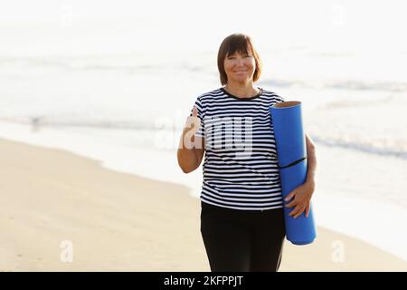 Frau im mittleren Alter, die eine Sportmatte hält und sich auf Yoga am Strand vorbereitet. Glückliche reife übergewichtige Frau, die am Meer trainiert Stockfoto