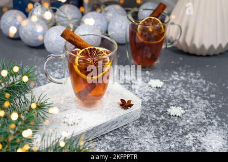 Zwei Tassen Tee aus Glas mit Zitronen- und Zimtstangen auf dem Hintergrund von Weihnachtsgirlanden und einem Geschenk. Stockfoto