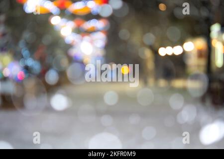 Abstrakter unscharfer Hintergrund. Menschen, die in der Winternacht während des starken Schneefalls auf der Straße mit geschmückten Weihnachtsbäumen, die mit leuchtenden Lichtern, glühenden Lichtern und Girlanden geschmückt sind, spazieren gehen Stockfoto