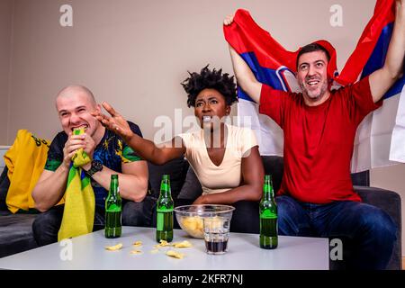 Freunde schauen sich ein Fußballspiel im Fernsehen an und jubeln ihre Nationalmannschaft an. Fußballweltmeisterschaft. Fans aus Brasilien gegen Fans aus Serbien. Stockfoto