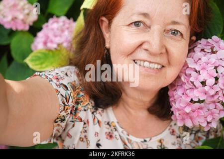 Selfie einer glücklichen reifen hispanischen Frau in einem Garten, umgeben von rosa Blumen, Hortensien. Konzepte: Einsatz von Technik, Genuss im Freien. Stockfoto