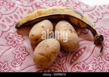 Ein reifer Kochbananen und drei weiße Kartoffeln stehen auf einer rot-weißen Tischdecke aus Spitze. Stockfoto