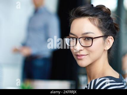 Sie strahlt Vertrauen in die Karriere aus. Porträt einer attraktiven jungen Frau, die in einem Büro mit Kollegen im Hintergrund sitzt. Stockfoto