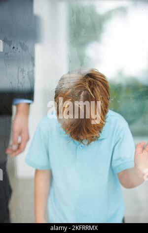 Ich wollte nach draußen gehen. Ein kleiner Junge, der enttäuscht mit seinem Kopf auf dem Fenster aussieht, als es regnet. Stockfoto