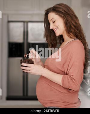 Dem Verlangen nach der Schwangerschaft nachgeben. Eine junge Schwangerin, die in der Küche einen Cupcake isst. Stockfoto