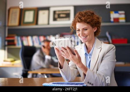 Online-Recherche. Eine junge Assistentin, die ein Tablet benutzt und ihren Chef im Hintergrund arbeitet. Stockfoto