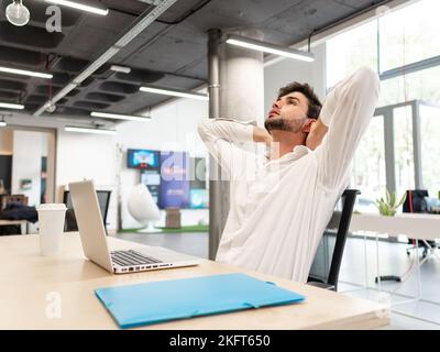 Erschöpfter bärtiger junger Mann in weißem Hemd, der mit Laptop und Ordner am Tisch sitzt und den Kopf in einem modernen weißen Büro nach hinten kippt Stockfoto