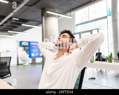 Erschöpfter bärtiger junger Mann in weißem Hemd sitzt am Tisch, während er den Kopf in einem modernen weißen Büro nach hinten kippt Stockfoto
