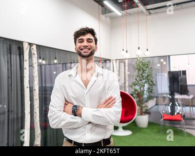 Lächelnder junger bärtiger Mann in weißem Hemd und Armbanduhr mit gekreuzten Armen, der in einem modernen Arbeitsbereich mit Glaswand steht und die Kamera anschaut Stockfoto