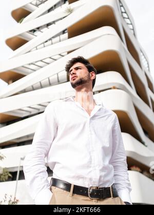 Junger bärtiger Mann mit dunklen Haaren in weißem Hemd, der auf der Straße und in einem modernen mehrstöckigen Gebäude steht und wegschaut Stockfoto