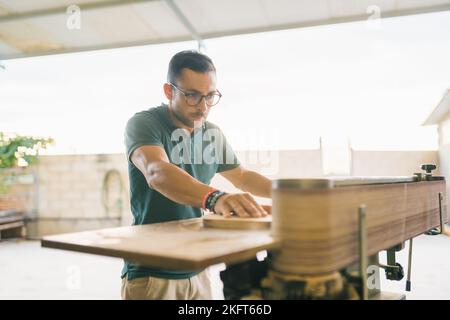 Seitenansicht des männlichen Zimmermanns in legerer Kleidung und Brille mit Bandschleifmaschine auf Holzbrett in der Werkstatt Stockfoto