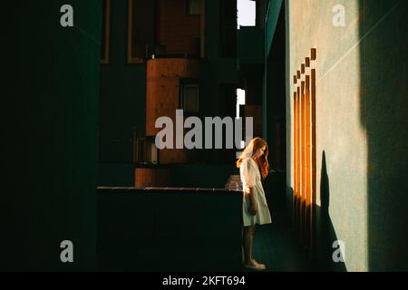 Seitenansicht einer Rothaarige Dame in weißem Kleid, die im dunklen Flur eines modernen Gebäudes in Barcelona steht, während die Sonne durch das Fenster nach unten scheint Stockfoto