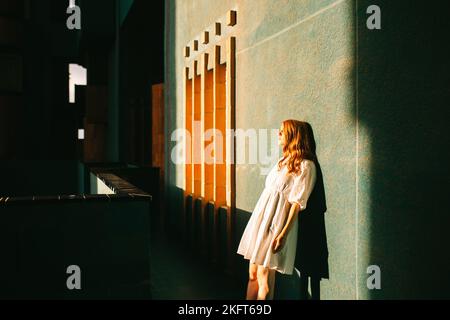 Seitenansicht einer Rothaarige Dame in weißem Kleid, die in einem dunklen Flur eines modernen Gebäudes in Barcelona steht, während die Sonne durch das Fenster scheint Stockfoto