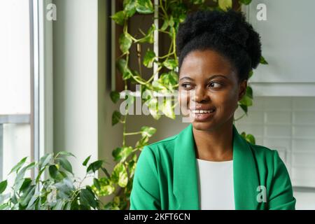 Fröhliche, fröhliche junge afrikanische Frau, die mit einem Lächeln aus dem Fenster schaut. Positives Wohlbefinden Stockfoto