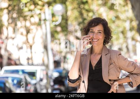 Lächelnde Geschäftsfrau mit lockigen Haaren, die auf dem Mobiltelefon spricht und auf der Straße der Stadt wegschaut Stockfoto