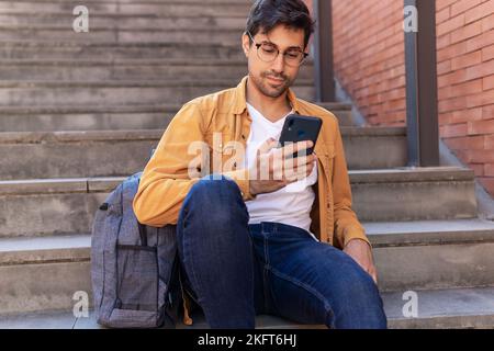 Fokussierter junger bärtiger hispanischer Mann in Jeans und Brillen, der auf einer Steintreppe mit Rucksack sitzt und tagsüber über das Mobiltelefon im Internet surft Stockfoto