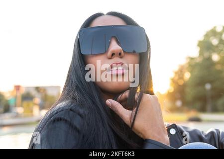 Junge hispanische Hündin mit stylischer Sonnenbrille, die das Kinn berührt und auf den verschwommenen Hintergrund von Sonnenuntergang am Himmel und im Park blickt Stockfoto
