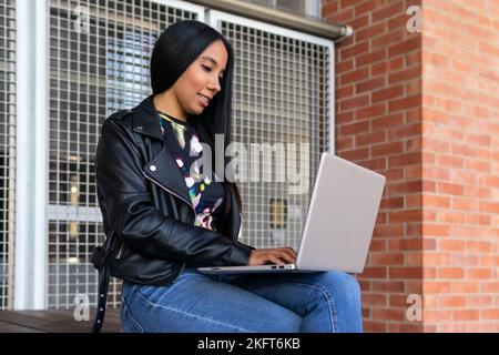 Junge hispanische Frau in legerer Kleidung lächelt und macht Hausaufgaben auf Netbook, während sie in der Nähe des Fensters und der Backsteinmauer des Universitätsgebäudes sitzt Stockfoto