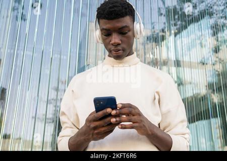 Niedriger Winkel des jungen afroamerikanischen Mannes in legerer Kleidung, der Musik in Kopfhörern hört und im Mobiltelefon surft, während er gegen den modernen BU steht Stockfoto