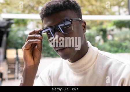 Ernsthafter afroamerikanischer Mann in eleganter Freizeitkleidung mit Brillen und Blick auf die Kamera vor verschwommenem Hintergrund Stockfoto