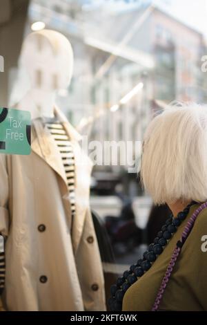 Seitenansicht einer anonymen älteren Dame mit kurzen Haaren, die in der Nähe der Vitrine stand und während des Spazierens auf der Stadtstraße eine Jacke ansah Stockfoto