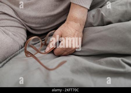 Von oben der Ernte anonyme ältere Frau in warmen Pullover sitzt auf dem Bett mit grauen Bettwäsche und hält Brillen Stockfoto