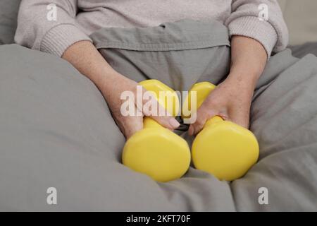 Hoher Winkel der Ernte anonyme Frau in grau warmer Pullover sitzt auf weichem Bett unter Decke und hält gelbe Hanteln Stockfoto