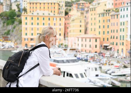 Seitenansicht einer erwachsenen Frau mit Rucksack, der auf einem Damm steht und vor unscharfem Hintergrund die Küstenstadt in Italien betrachtet Stockfoto