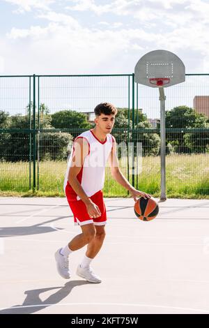 Entschlossener kaukasischer männlicher Streetball-Spieler, der im Sommer auf dem Sportplatz Basketball spielt Stockfoto