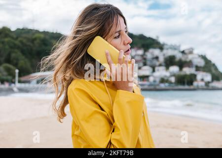 Seitenansicht der Crop-Hündin mit langen blonden Haaren in gelbem Regenmantel, die mit ihrem Smartphone auf dem verschwommenen Hintergrund der grünen Insel Spanien anruft Stockfoto