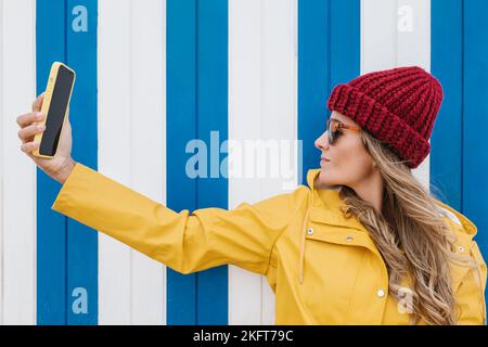 Seitenansicht einer Crop-Hündin mit langen blonden Haaren in gelbem Regenmantel, die ein Selfie mit ihrem Smartphone in der Nähe eines gestreiften Gebäudes machte Stockfoto