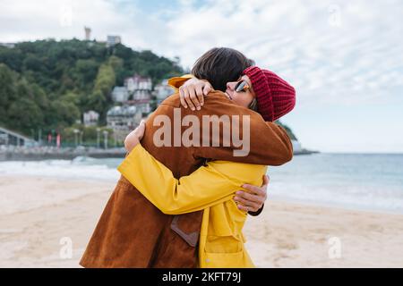 Seitenansicht einer fröhlichen Frau, die lächelt und einen Mann umarmt, beide in Jacken, während sie am malerischen Strand von Ondarreta in Donostia in Spai steht Stockfoto