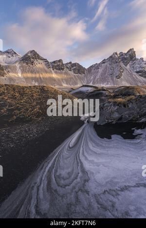 Ruhiges, gefrorenes, endloses Meer, umgeben von schwarzem, gefrorenem Sand und hohem, schneebedeckten Berg Vestrahorn bei einem malerischen Sonnenuntergang am Stockness Beach, Icel Stockfoto